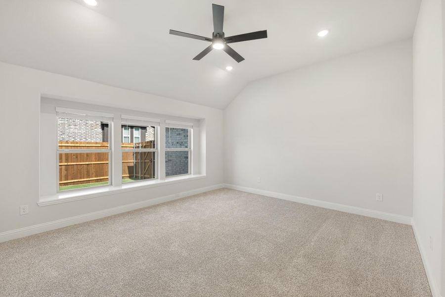 Empty bedroom with vaulted ceiling, ceiling fan, carpet flooring, and triple window overlooking backyard fence