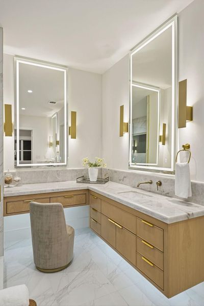 The sprawling "her" vanity serves as a dedicated dressing and beauty station, featuring an expansive L-shaped marble countertop with a built-in makeup desk and plush seating nook. Twin backlit mirrors and architectural gold lighting fixtures frame the space, creating a gallery-like atmosphere.