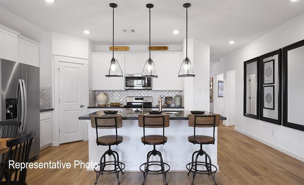 Modern kitchen featuring white cabinetry, stainless steel appliances, a herringbone tile backsplash, an island with dark countertops, and wood-finish flooring Modern kitchen featuring white cabinetry, stainless steel appliances, a herringbone tile backsplash, an island with dark countertops, and wood-finish flooring