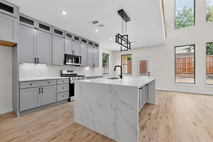 Kitchen with gray cabinetry, backsplash, appliances with stainless steel finishes, hanging light fixtures, and light stone countertops Kitchen with gray cabinetry, backsplash, appliances with stainless steel finishes, hanging light fixtures, and light stone countertops