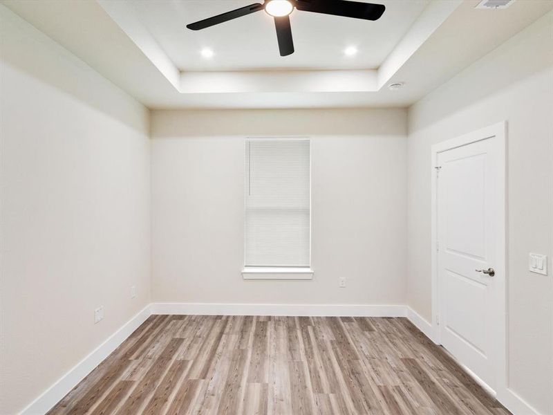 Room with wood-finish flooring, a tray ceiling with recessed lighting, a ceiling fan, a window with blinds, and a paneled door Room with wood-finish flooring, a tray ceiling with recessed lighting, a ceiling fan, a window with blinds, and a paneled door