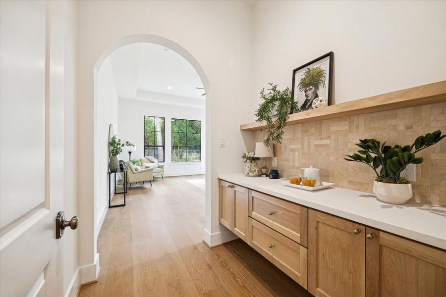 Hallway wet bar with stone backsplash, wood cabinetry, open shelf, and arched entry to the primary suite.