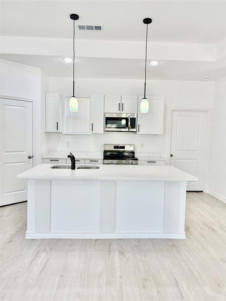 Kitchen featuring white cabinetry, hanging light fixtures, an island with sink, stainless steel appliances, and light stone countertops