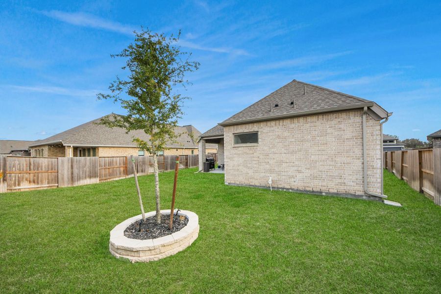 Spacious backyard with a well-maintained lawn, a young tree in a stone-bordered planter, and privacy fencing surrounding a brick exterior home. Spacious backyard with a well-maintained lawn, a young tree in a stone-bordered planter, and privacy fencing surrounding a brick exterior home.