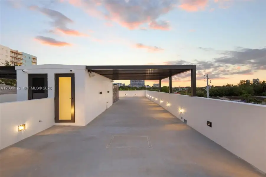 Rooftop terrace featuring smooth concrete flooring, solid perimeter walls, built-in wall-mounted lighting, and a metal-framed covered area Rooftop terrace featuring smooth concrete flooring, solid perimeter walls, built-in wall-mounted lighting, and a metal-framed covered area