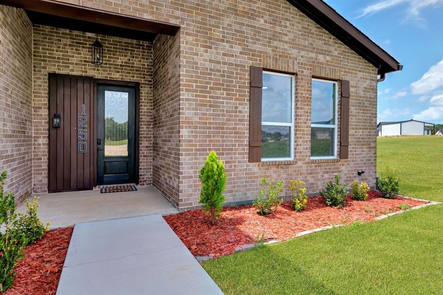 Brick exterior with a dark-framed glass paneled front door, wood-finish shutters, and a concrete pathway Brick exterior with a dark-framed glass paneled front door, wood-finish shutters, and a concrete pathway