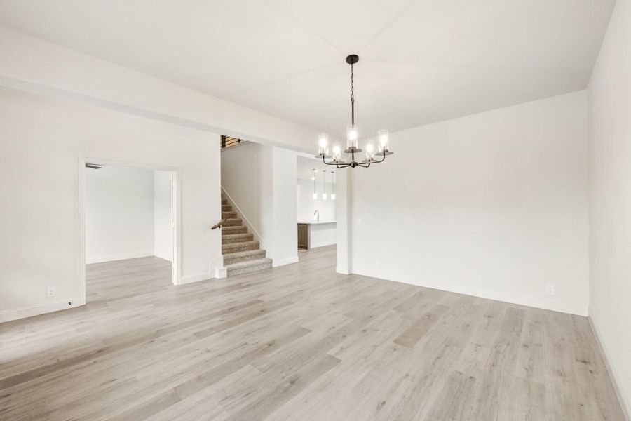 Open living and dining area with light hardwood floors, chandelier, and staircase visible in background Open living and dining area with light hardwood floors, chandelier, and staircase visible in background