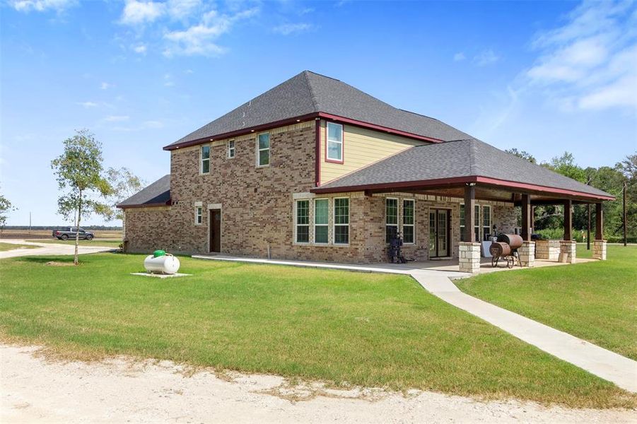 This photo showcases a spacious two-story brick home with a large wrap-around porch, set in a well-maintained grassy yard. The house features a modern roof and ample outdoor space, ideal for relaxation or entertaining. The property has a rural feel, with open skies and a peaceful setting.