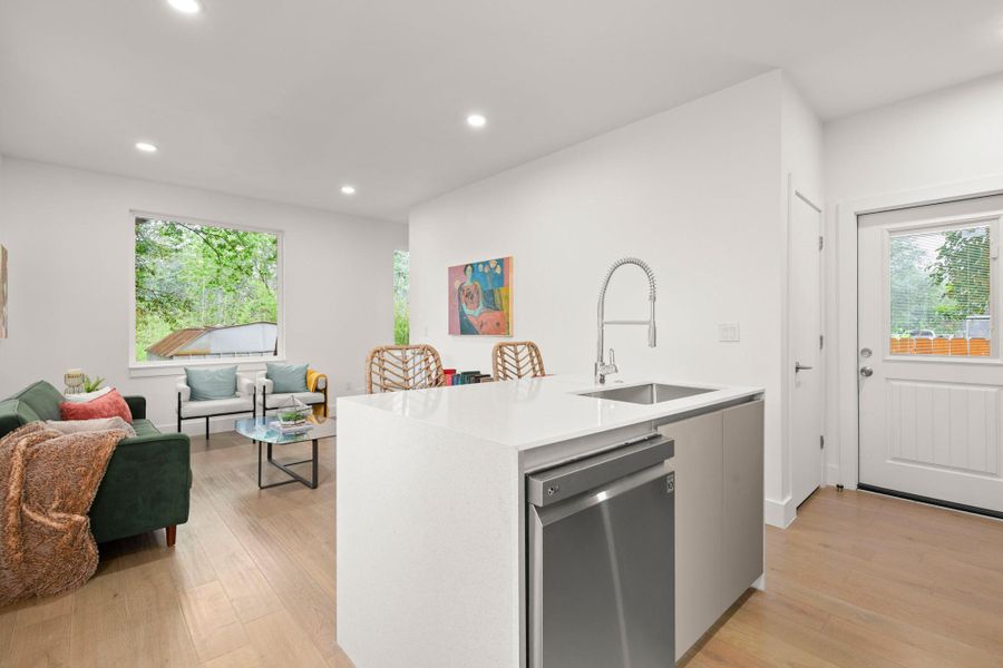 Open-concept living area featuring light wood-finish flooring, recessed ceiling lighting, and a kitchen island with a built-in stainless steel sink and appliance