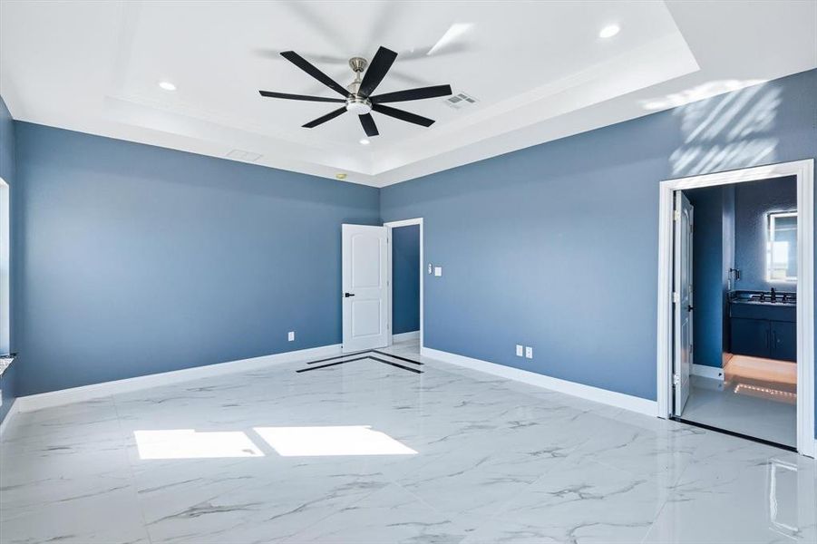 Unfurnished bedroom with a raised ceiling, light marble finish floors, a ceiling fan, recessed lighting, and ensuite bath