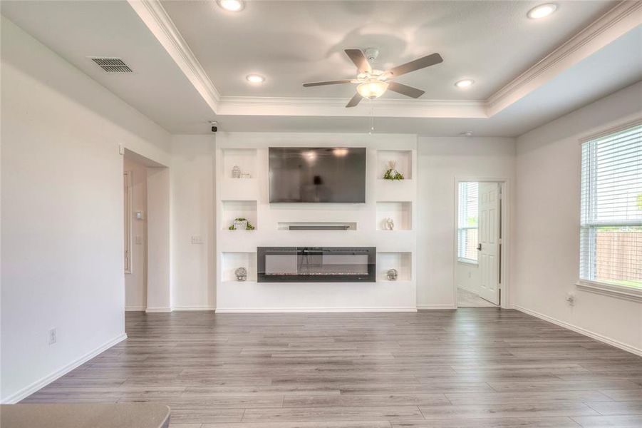 Unfurnished living room with a glass covered fireplace, wood finished floors, a ceiling fan, crown molding, and a raised ceiling Unfurnished living room with a glass covered fireplace, wood finished floors, a ceiling fan, crown molding, and a raised ceiling