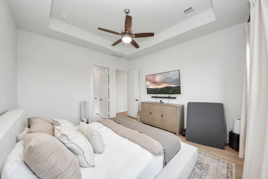 Spacious bedroom with a tray ceiling, ceiling fan, and natural light. Features a cozy bed, wall-mounted TV, and elegant wood cabinetry.
