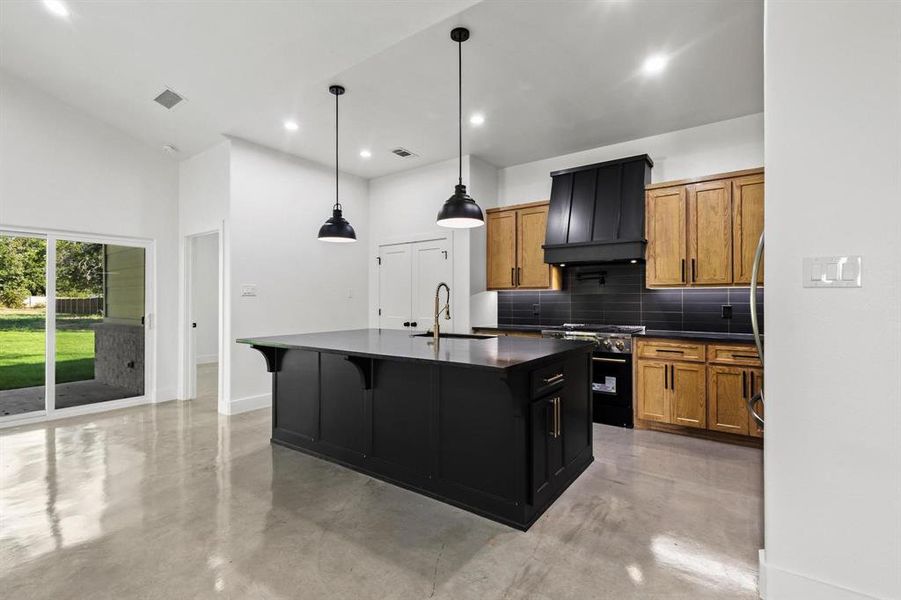 Kitchen with dark cabinetry, decorative backsplash, an island with sink, gas range, and finished concrete flooring