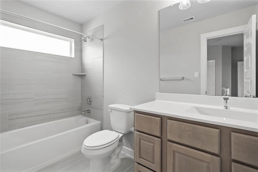 Secondary bath features tile flooring, bath/shower combo with tile surround, wood stained cabinets, beautiful light countertops, mirror, dark, sleek fixtures and modern finishes!