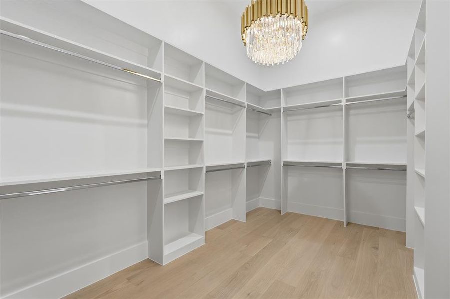 Walk-in closet featuring built-in white shelving, chrome hanging rods, wood-finish flooring, and a gold-tone chandelier with crystal accents Walk-in closet featuring built-in white shelving, chrome hanging rods, wood-finish flooring, and a gold-tone chandelier with crystal accents