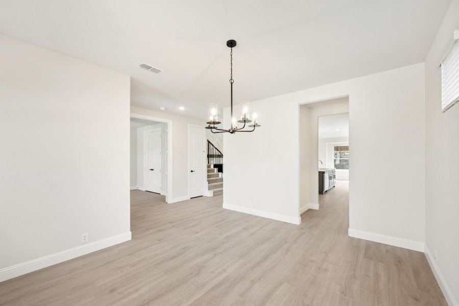 Empty dining room with light wood floors, white walls, black chandelier, and open view to kitchen and staircase
