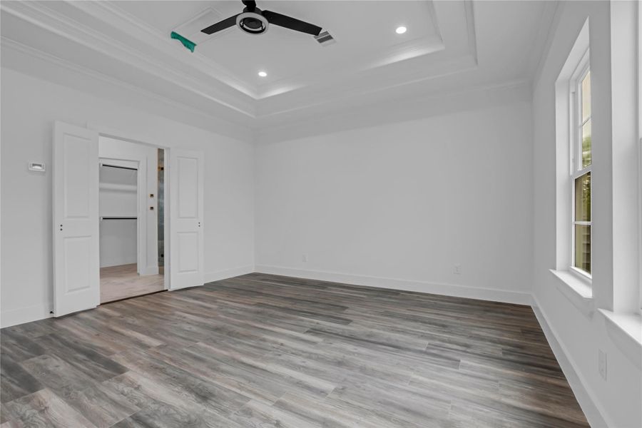 This spacious bedroom boasts elegant tray ceilings, sleek wood flooring, and ample natural light, offering a tranquil retreat with an attached walk-in closet for ultimate convenience.