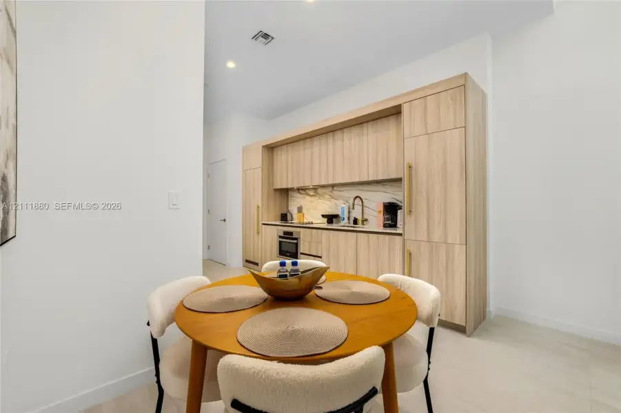 Kitchenette featuring light wood-finish cabinetry with integrated appliances, stone-look backsplash, recessed lighting, and a gold-tone faucet