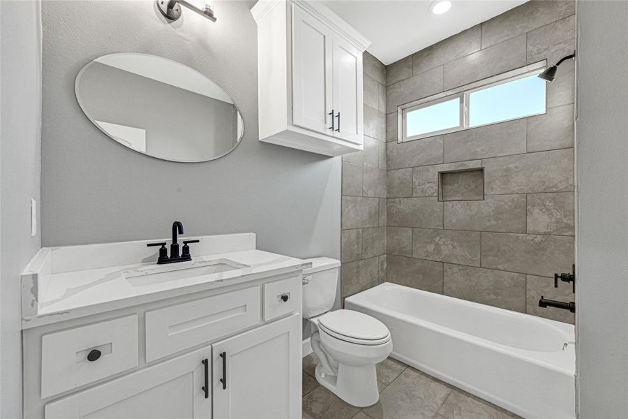 This modern hall  bathroom features a sleek design with a white vanity and marble countertop, a rectangular mirror, and elegant black fixtures. It includes a bathtub with a tiled shower, a built-in niche, and a frosted window for natural light. The space is enhanced by neutral gray walls and ample storage with a mounted cabinet.