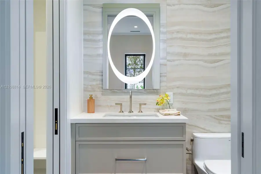 Custom vanity with stone-surface countertop, integrated rectangular sink, oval backlit mirror, and floor-to-ceiling stone-tiled wall Custom vanity with stone-surface countertop, integrated rectangular sink, oval backlit mirror, and floor-to-ceiling stone-tiled wall