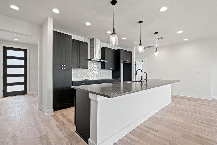 Kitchen featuring dark cabinetry, wall chimney exhaust hood, an island with sink, decorative light fixtures, and light wood-style floors