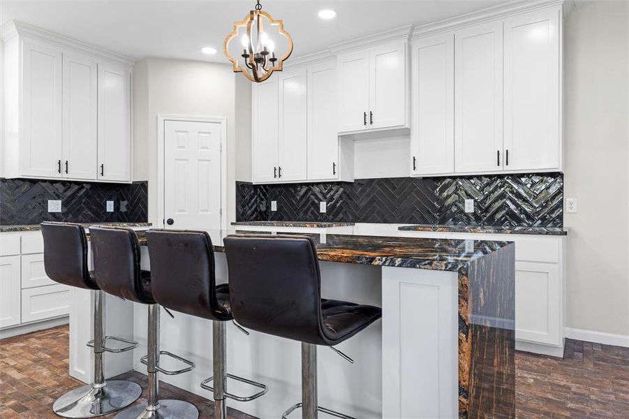 Kitchen featuring dark stone counters, decorative backsplash, white cabinets, a kitchen bar, and hanging light fixtures Kitchen featuring dark stone counters, decorative backsplash, white cabinets, a kitchen bar, and hanging light fixtures