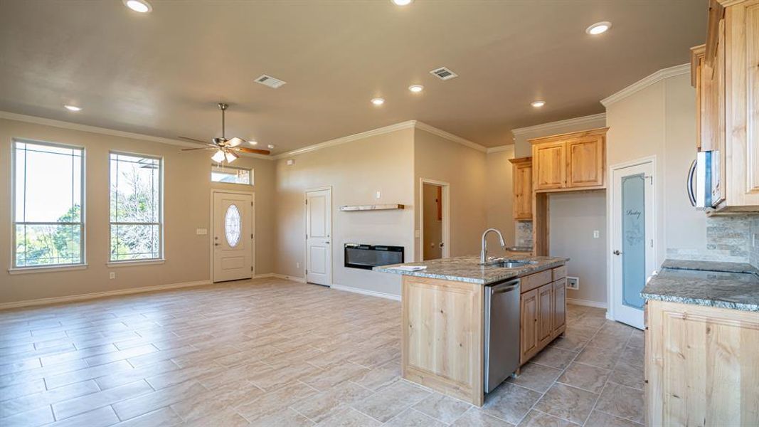 Kitchen with light brown cabinets, light stone counters, crown molding, ceiling fan, and a center island with sink