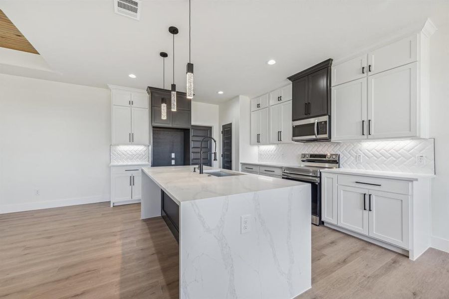 Kitchen with appliances with stainless steel finishes, backsplash, light hardwood / wood-style flooring, hanging light fixtures, and an island with sink