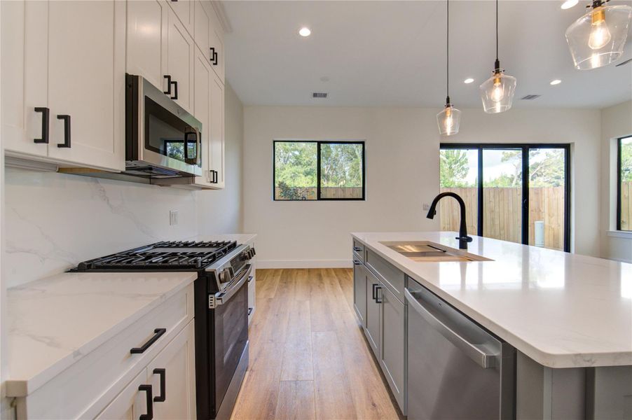 This modern kitchen features sleek white cabinetry, a spacious island with a built-in sink, and stainless steel appliances. The room is brightened by pendant lighting and large windows, offering a view of the outside. The vinyl plank flooring adds warmth and complements the contemporary design. This modern kitchen features sleek white cabinetry, a spacious island with a built-in sink, and stainless steel appliances. The room is brightened by pendant lighting and large windows, offering a view of the outside. The vinyl plank flooring adds warmth and complements the contemporary design.