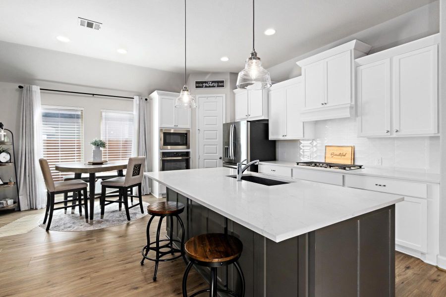 This modern kitchen features sleek white cabinetry, a large island with a sink and seating, stainless steel appliances, and stylish pendant lighting. Adjacent is a cozy dining area with natural light from two windows, creating a bright and inviting space.