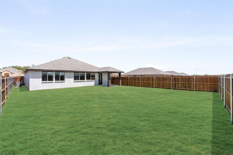 Backyard with large green lawn, wood privacy fence, and single-story brick home with covered patio