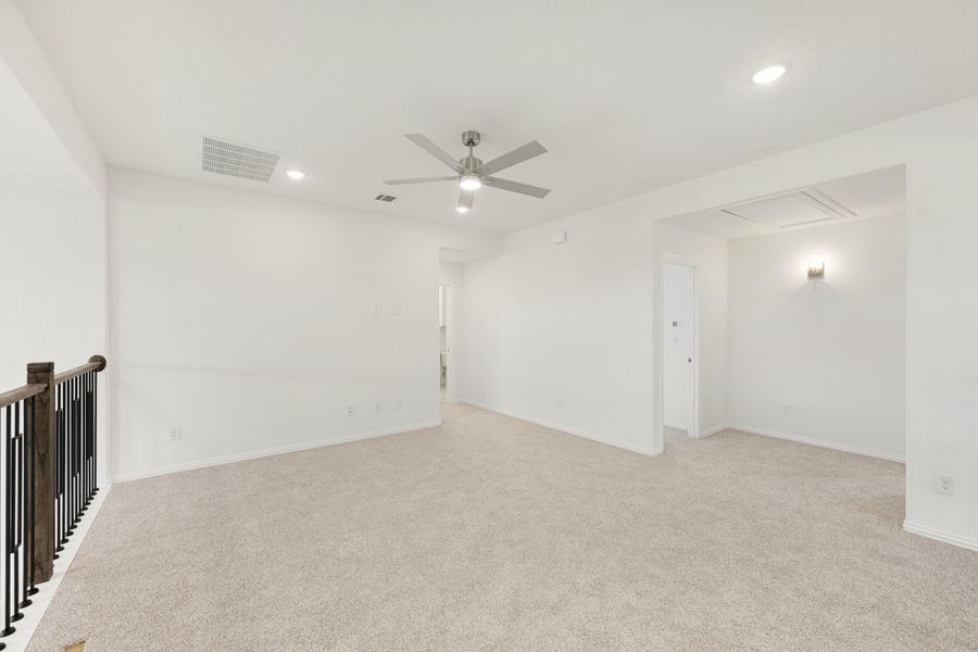 Empty upstairs loft with beige carpet, ceiling fan, and staircase railing visible in the foreground