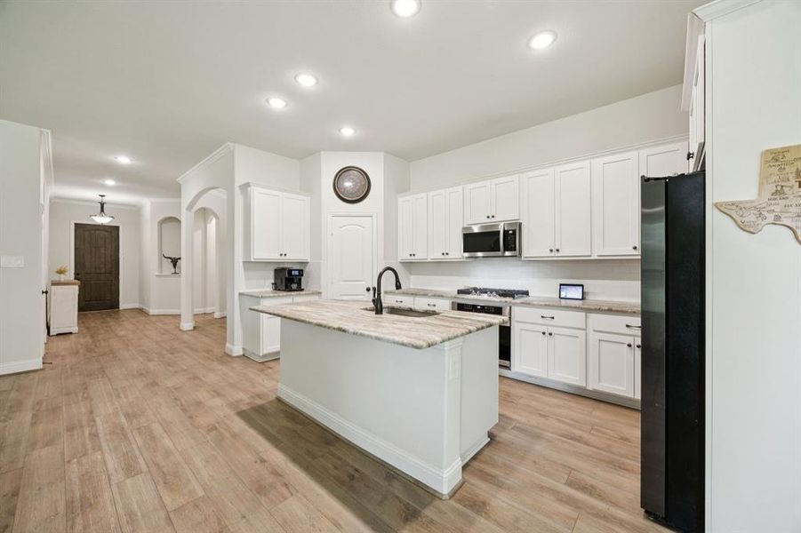 Open concept kitchen featuring white cabinets, granite counter tops, stainless steel appliances, recessed lighting, vinyl plank floors floors, and an island with sink.