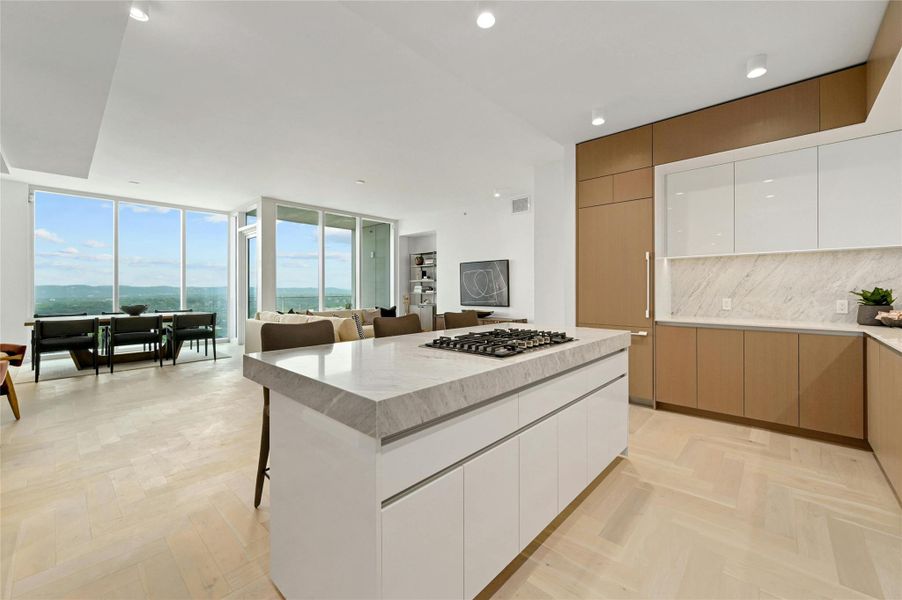 A modern, open-concept kitchen, dining and living area. The kitchen features a sleek, minimalist design with a large island at its center, topped with white-colored stone countertop. The island includes a built-in gas cooktop. Custom cabinetry seamlessly conceals the integrated refrigerator, maintaining clean lines and a refined aesthetic.