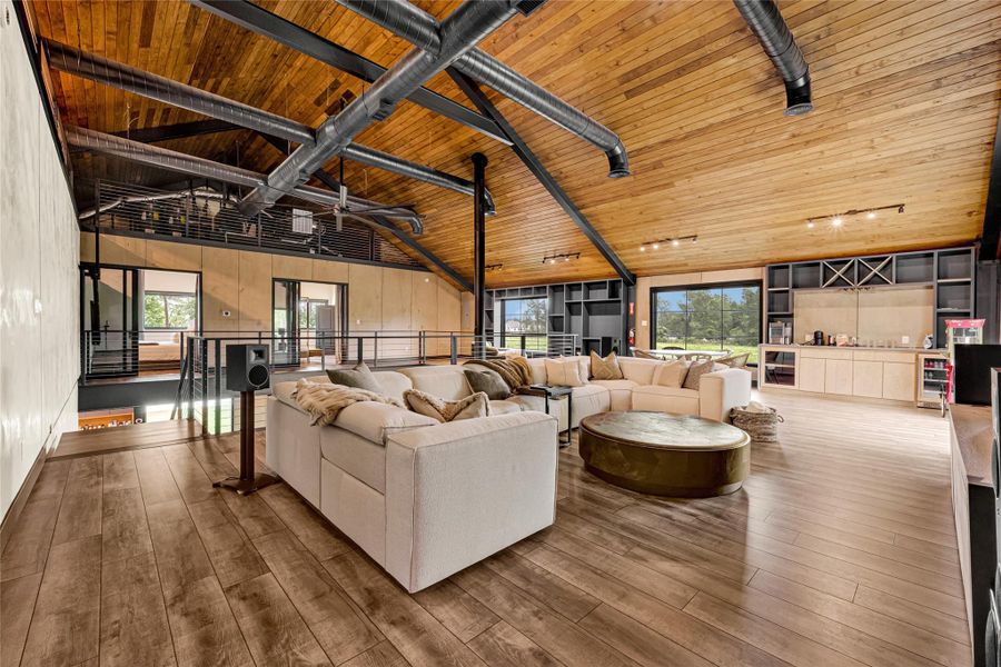 High wood-paneled ceilings and exposed ductwork. The room is open-concept, with a cozy sectional sofa, large windows offering natural light, and a built-in entertainment center. A sleek bar area adds functionality and style, perfect for entertaining.