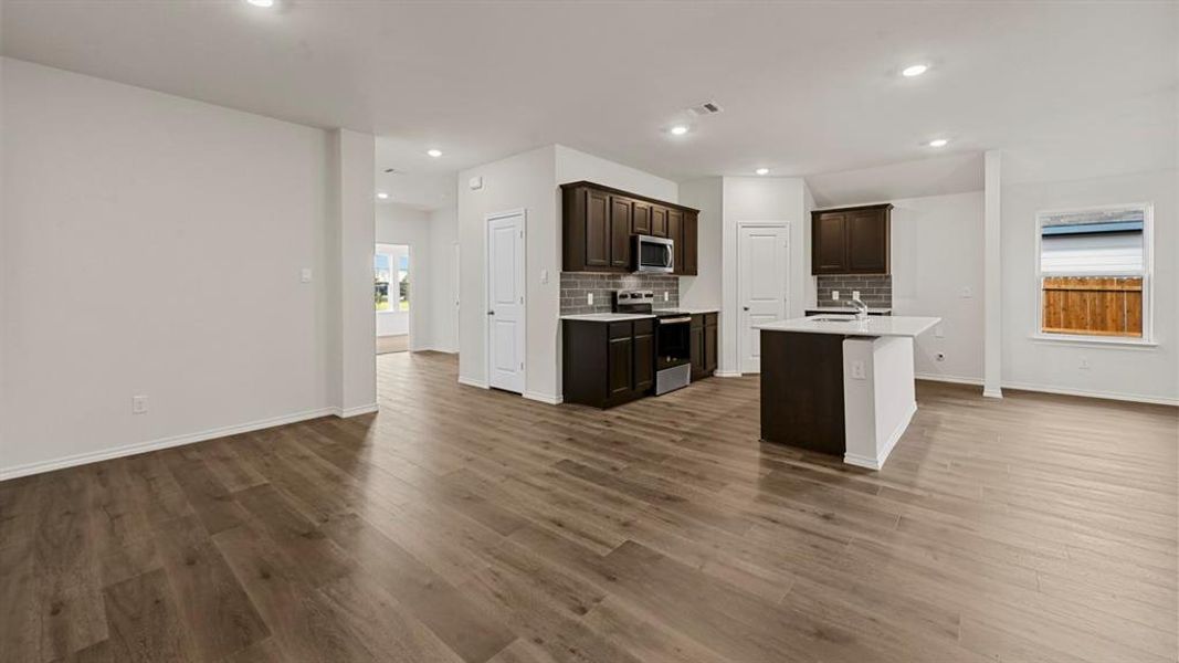 Open-concept kitchen featuring wood-finish flooring, dark wood cabinetry, stainless steel appliances, a white island with an integrated sink, and recessed lighting Open-concept kitchen featuring wood-finish flooring, dark wood cabinetry, stainless steel appliances, a white island with an integrated sink, and recessed lighting
