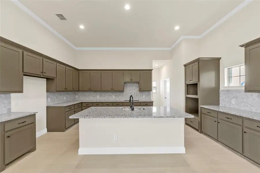 Kitchen with decorative backsplash, light stone counters, a kitchen island with sink, crown molding, and gray cabinetry Kitchen with decorative backsplash, light stone counters, a kitchen island with sink, crown molding, and gray cabinetry