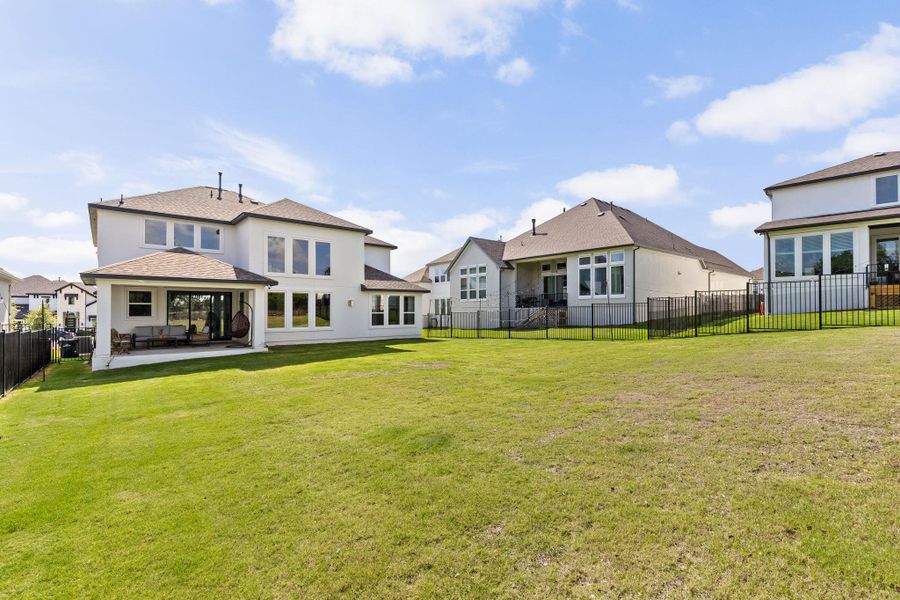 Expansive backyard featuring a manicured lawn, a covered patio with a concrete slab, multiple large windows, and a dark shingle roof