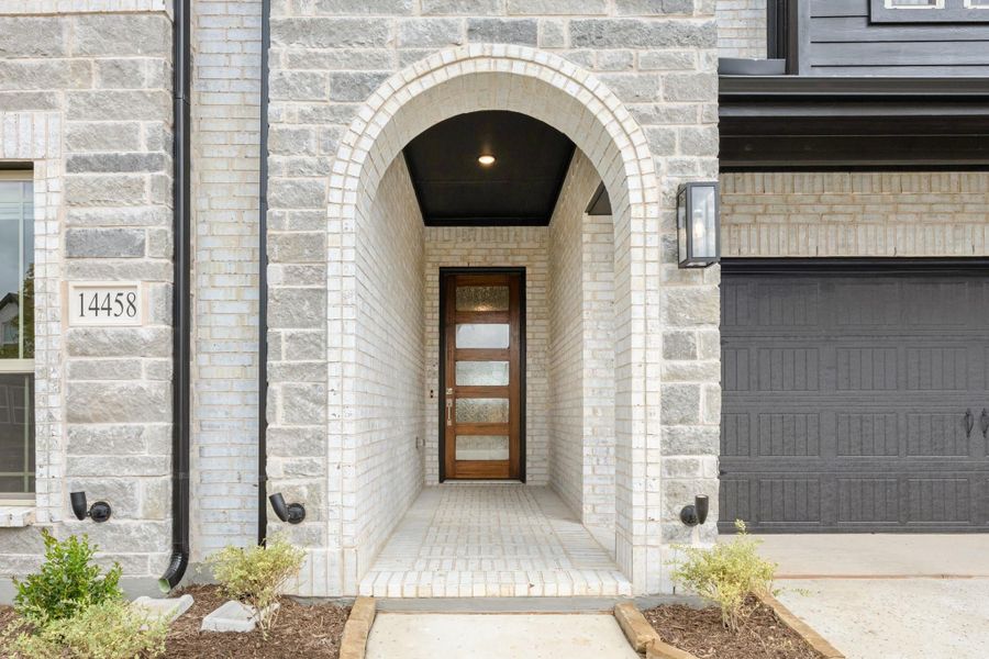 Stone archway entrance with wood and glass front door, brick walkway, and dark garage door beside it