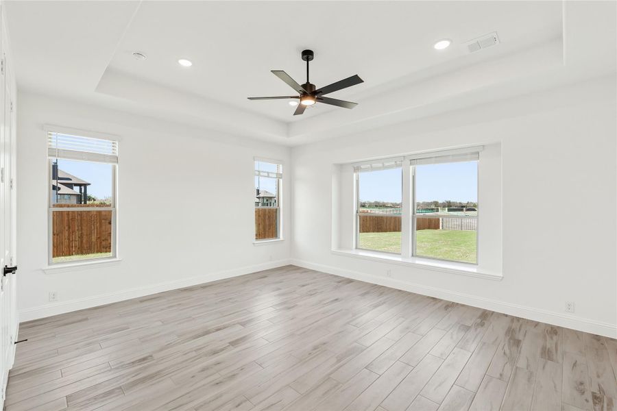 Empty room with light wood floors, ceiling fan, tray ceiling, and large windows overlooking a backyard Empty room with light wood floors, ceiling fan, tray ceiling, and large windows overlooking a backyard