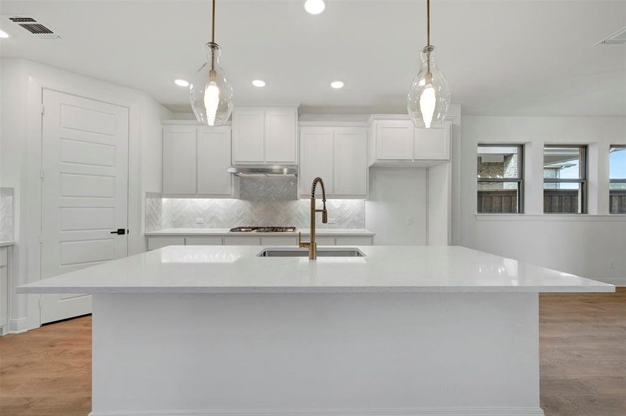 Kitchen featuring light wood-style floors, tasteful backsplash, a kitchen island with sink, white cabinetry, and light stone counters
