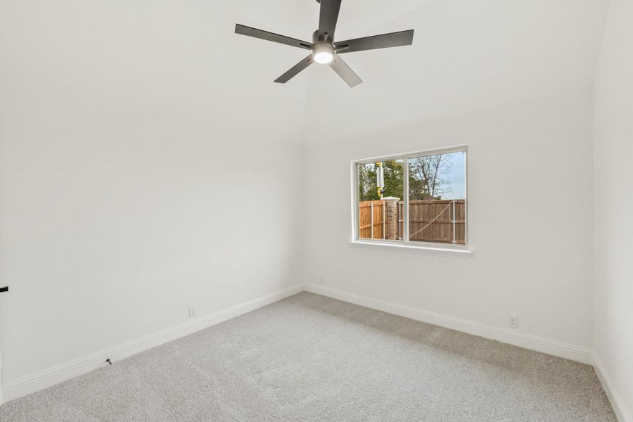 Empty bedroom with gray carpet, vaulted ceiling, ceiling fan, and single window overlooking fenced backyard