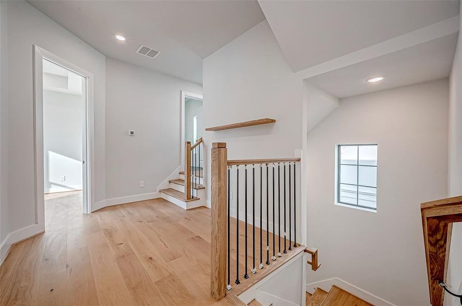 This photo showcases a the third floor. The landing is bright, modern with light wood flooring and a staircase featuring wooden handrails and black spindles. The space is well-lit with recessed lighting and has a window providing natural light. The primary bedroom and a third bedroom is on this floor.