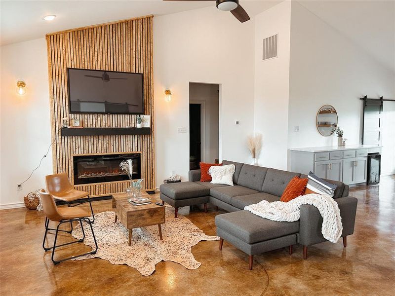 Living area featuring finished concrete floors, a barn door, high vaulted ceiling, a glass covered fireplace, and a ceiling fan Living area featuring finished concrete floors, a barn door, high vaulted ceiling, a glass covered fireplace, and a ceiling fan