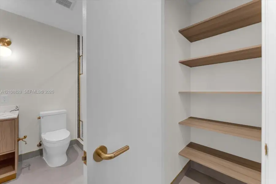 Built-in storage closet featuring tiered wood-finish shelving, brass hardware, neutral-toned cabinetry, and minimalist wall-mounted lighting
