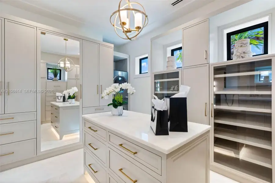 Custom built-in cabinetry with integrated lighting, central island with stone countertop, full-length mirror, glass-front display shelving, and orb-style metallic pendant light fixture Custom built-in cabinetry with integrated lighting, central island with stone countertop, full-length mirror, glass-front display shelving, and orb-style metallic pendant light fixture
