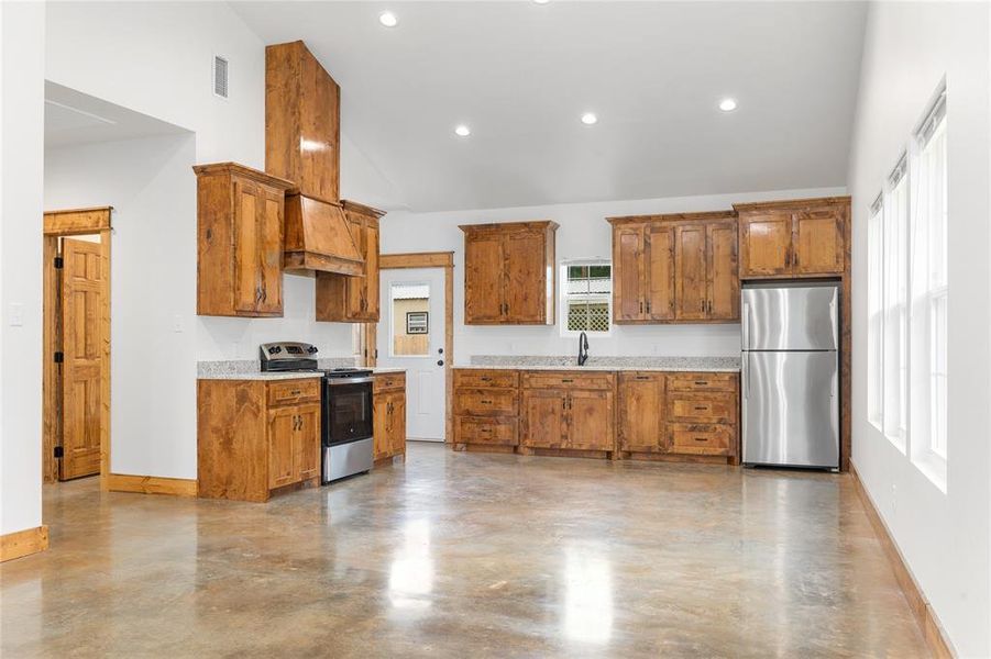 Kitchen with appliances with stainless steel finishes, concrete flooring, custom exhaust hood, brown cabinets, and high vaulted ceiling Kitchen with appliances with stainless steel finishes, concrete flooring, custom exhaust hood, brown cabinets, and high vaulted ceiling