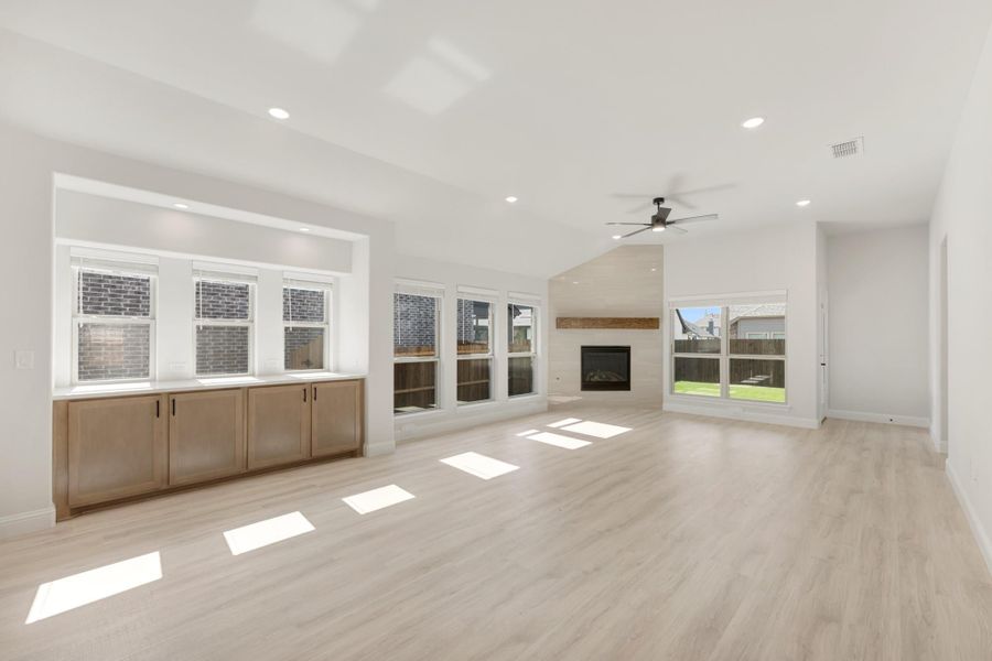 Open living room with light wood floors, fireplace, ceiling fan, and built-in cabinets below large windows