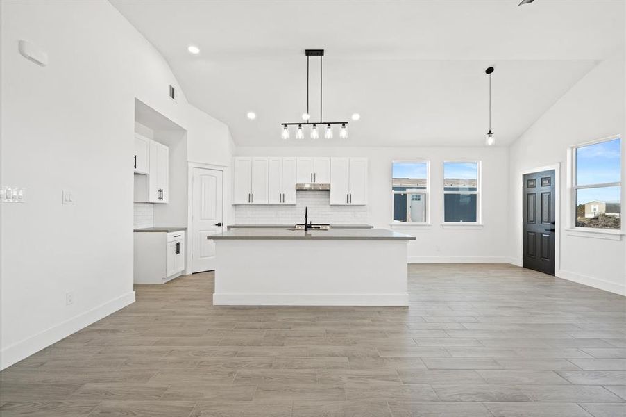 Spacious, modern kitchen with large center island, white cabinetry, sleek countertops, and stainless steel fixtures. Natural light fills the room from multiple windows. Contemporary lighting fixtures add style to the clean, bright space.