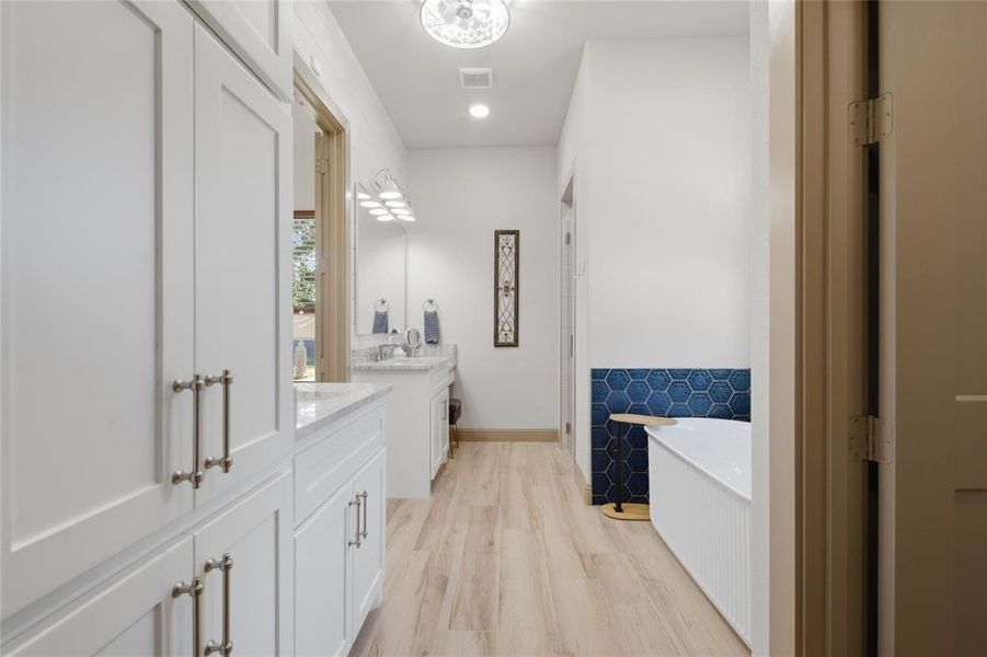 Bright bathroom featuring extensive white cabinetry with brushed nickel hardware, a dual vanity with a light-toned countertop, a soaking tub with blue hexagonal tile surround, and wood-finish flooring